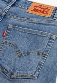 Jeans en denim avec un délavage bleu clair, dotés de coutures jaune doré, d'une poche arrière au design incurvé distinctif et d'une étiquette en cuir beige.