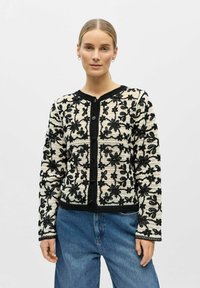 Femme portant un cardigan blanc en tricot avec une broderie florale noire et un jean bleu, debout devant un fond clair uni.