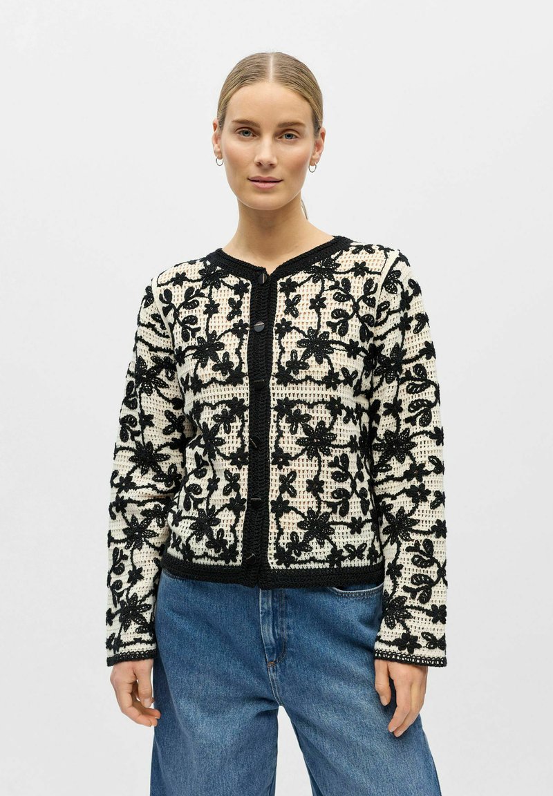 Femme portant un cardigan blanc en tricot avec une broderie florale noire et un jean bleu, debout devant un fond clair uni.