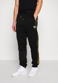 Svarta atletiska joggers med guld sidan ränder, elastisk midjeband med snörning, med en Adidas-logotyp på vänster höft. Vita sneakers.