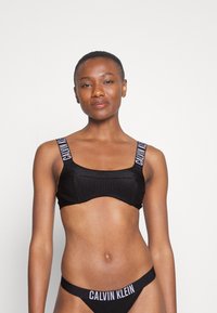 Črn rebrast bralette s kvadratnim izrezom in širokimi naramnicami, ki vsebuje logotip "CALVIN KLEIN". Ujemajoči črn tangice z logotipom na pasu.
