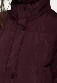 Burgundská zateplená bunda s vysokým límcem, zapínáním na přední cvočky a prošívaným designem s hladkou, lesklou texturou látky.