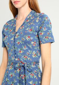 Blommig klänning med blå bakgrund, korta ärmar, V-ringning, knapprad fram och en knytbelte i midjan. Innehåller en mängd färgglada blommor.