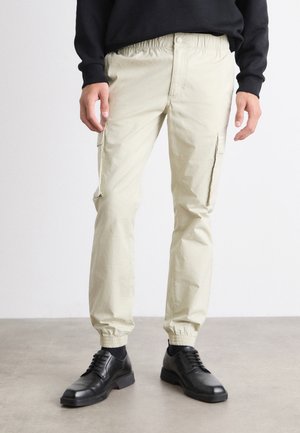 Personne portant un pantalon cargo beige avec des poignets élastiques et des chaussures habillées noires, debout sur un sol gris devant un fond blanc.