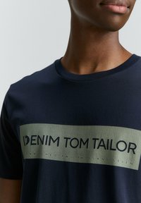 Kara jūras zila kokvilnas T-krekls ar taisnstūrveida olīvu zaļu grafiku, kurā ir uzraksts "DENIM TOM TAILOR" un punktiņu raksts. Apaļš kakla izgriezums.