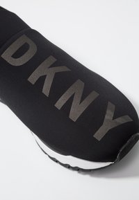 Chaussure de sport noire avec un upper de type chaussette arborant un grand logo DKNY réfléchissant. La semelle est blanche avec des détails de traction noirs.