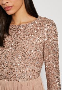 Abito beige con maniche lunghe, decorato con paillettes e perline. Fondo plissettato che aggiunge texture. Design semplice con scollatura tonda.