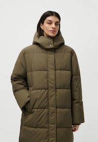 Woman with short dark hair wearing an oversized olive green puffer jacket with high collar and hands in pockets against white background.