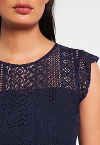 Top in pizzo blu navy con scollatura rotonda, caratterizzato da motivi intricati e maniche a volant, realizzato in tessuto leggero e testurizzato.