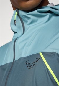 Jachetă sport ușoară în nuanțe de teal și gri, având un logo pe piept, accente galbene și o glugă ajustată. Textură netedă.
