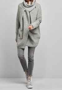 Ljust grå oversized poncho med en cowl-neck, stickad textur och en sned dragkedja. Matchad med grå jeans och ljusgrå sneakers.