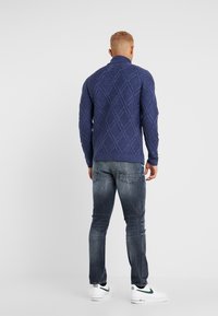 Maglione blu lavorato a maglia con motivo a rombi, colletto alto e polsini a coste, abbinato a jeans blu scoloriti e sneakers bianche.