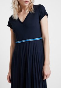 Mörkblå klänning med V-ringning och korta ärmar. Har en plisserad kjol och ett horisontellt randigt dekorativt band i blått och vitt vid midjan.