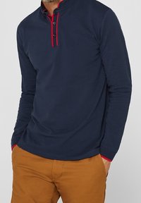 Mörkblå långärmad polo med hög krage, röda kontrastsömmar och placket med två knappar; bärs med beige byxor.