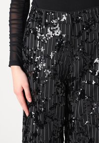 Pantaloni neri con paillettes, caratterizzati da strisce verticali, vita aderente e una superficie testurizzata con una combinazione di paillettes nere lucide e argento.