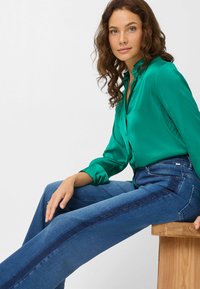 Groene satijnen overhemd met knoopjes, gecombineerd met blauwe wijde jeans met een donkerblauwe zijpaneel en subtiele versieringen.