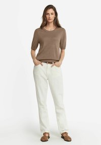 Brun kortærmet sweater, afslappet pasform; parret med hvide vide bukser; brune sandaler med vævet detalje; modellen står med hænderne i lommerne.