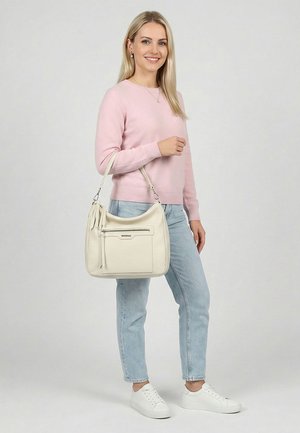 Vrouw draagt een roze trui en lichtblauwe spijkerbroek, houdt een gebroken witte schoudertas vast, staat tegen een effen lichte achtergrond en lacht.