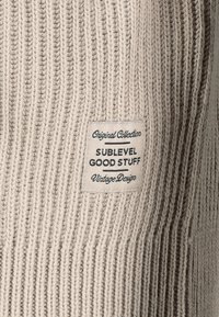 Beige gerippter Strickstoff mit einem gewebten Etikett, auf dem "Original Collection, SUBLEVEL, GOOD STUFF, Vintage Design" steht. Strukturiert Oberfläche und lässiger Stil.