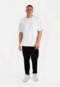 Witte katoenen t-shirt met korte mouwen en een kleine goudkleurige tekstdetail, gecombineerd met zwarte broek en witte sneakers. Simpele, casual look.