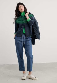 Granatowy sweter z dzianiny z zielonym wzorzystym karczkiem, nałożony na jasną zieloną golf, połączony z jasnoniebieskimi spodniami o szerokich nogawkach i białymi trampkami.