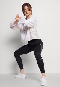 Weißer Sportjacke mit Reißverschluss und langen Ärmeln, kombiniert mit schwarzen Leggings, die drei weiße Streifenakzente aufweisen, und weißen Sportschuhen.