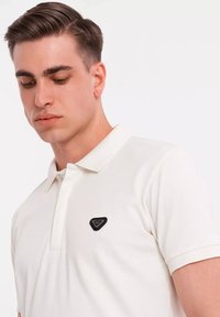 Hvid polo shirt med et sort trekantet logo på venstre bryst, klassisk krave og korte ærmer. Glat stofstruktur.