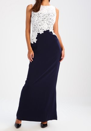 Kobieta w sukni bez rękawów sięgającej do ziemi, z białym koronkowym gorsetem z motywem kwiatowym i granatowym, gładkim dołem, w czarnych szpilkach z ostrym noskiem.