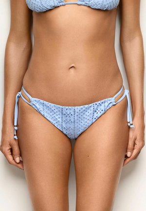 Kobieta nosząca jasnoniebieskie figi bikini z oczkami i wiązaniami po bokach, pokazująca środek ciała i ręce na neutralnym tle.