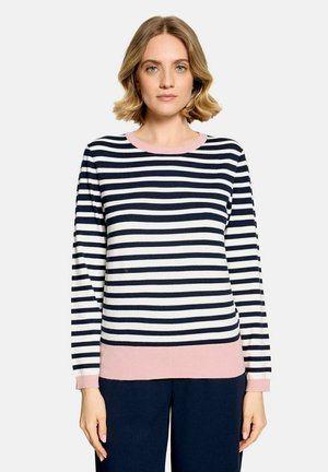 Femme aux cheveux blonds courts portant un pull à manches longues rayé noir et blanc avec une bordure rose et un pantalon foncé sur un fond uni.