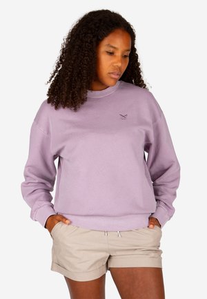 Frau mit lockigem Haar, die einen lavendelfarbenen Pullover und beige Shorts trägt, mit den Händen in den Taschen, schaut nach unten.