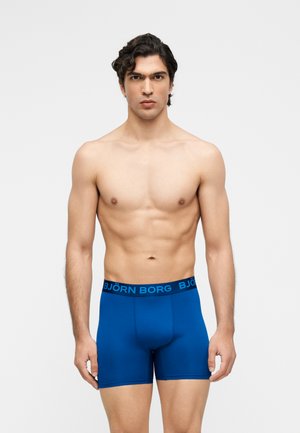 Jeune homme aux cheveux bouclés foncés, portant un boxer bleu Björn Borg, debout devant un fond uni gris clair, face à l'objectif.