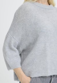 Femme portant un pull tricoté gris clair doux à manches trois-quarts, la main reposant dans la poche de son jean, fond neutre.