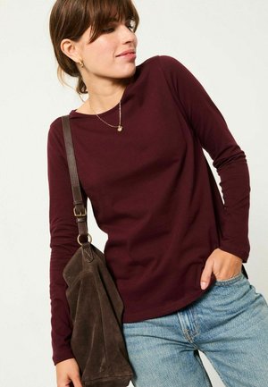 Burgunderfarbendes Langarmhemd aus weichem Material, kombiniert mit blauen Jeans und einer braunen Wildleder-Umhängetasche. Einfaches, lässiges Design.