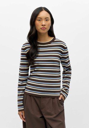 Femme aux longs cheveux foncés portant un haut rayé marron, bleu et blanc à manches longues et un pantalon marron ample, une main dans la poche.