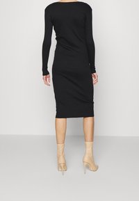 Robe midi noire à manches longues avec une silhouette ajustée, une texture lisse et un dos échancré. Portée avec des bottines à talons hauts couleur nude.