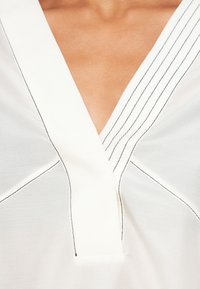 Hvit bluse med V-utringning, med kontrasterende svart søm langs sømmene og subtil tekstur. Glatt stoff, minimalistisk design.