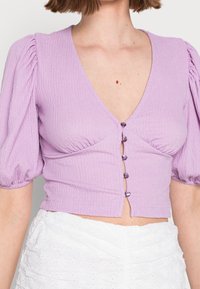Blusa di lavanda con texture, maniche a palloncino corte, scollatura a V profonda e sei bottoni viola sul davanti. Tessuto liscio con dettagli arricciati.