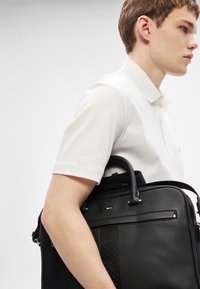 Μαύρη δερμάτινη τσάντα με υφασμάτινες λεπτομέρειες, δύο χερούλια και ιμάντα ώμου. Το μοντέλο που φοράει λευκό πόλο κρατά τη τσάντα.