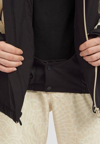 Zwarte jas met een zachte binnenkant, drukknoopsluiting bij de taille en een rits, gedragen over lichtgekleurde broek met textuur en een patroon.