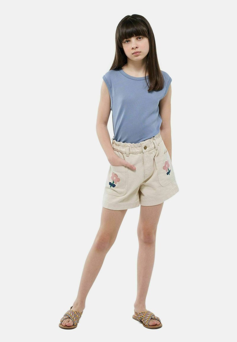 Haut sans manches bleu clair associé à un short beige avec des broderies florales sur les poches, une taille élastique et des sandales plates.