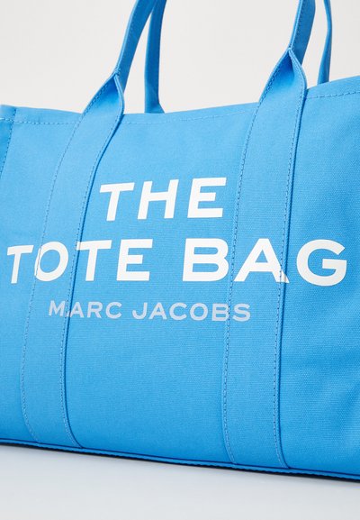 La borsa a tracolla blu in tela presenta manici robusti, un evidente testo bianco che recita "THE TOTE BAG" e "MARC JACOBS", con dettagli di cucitura visibili.