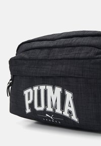 Černá sportovní taška vyrobená z texturovaného materiálu, která má výrazné bílé logo "PUMA" a decentní brandingové detaily. Zapínání na zip v horní části.
