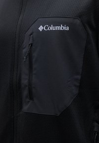 Πορτοκαλί μπουφάν με υφή στην περιοχή του θώρακα, με τσέπη με φερμουάρ και λευκό λογότυπο "Columbia" πάνω από την τσέπη.