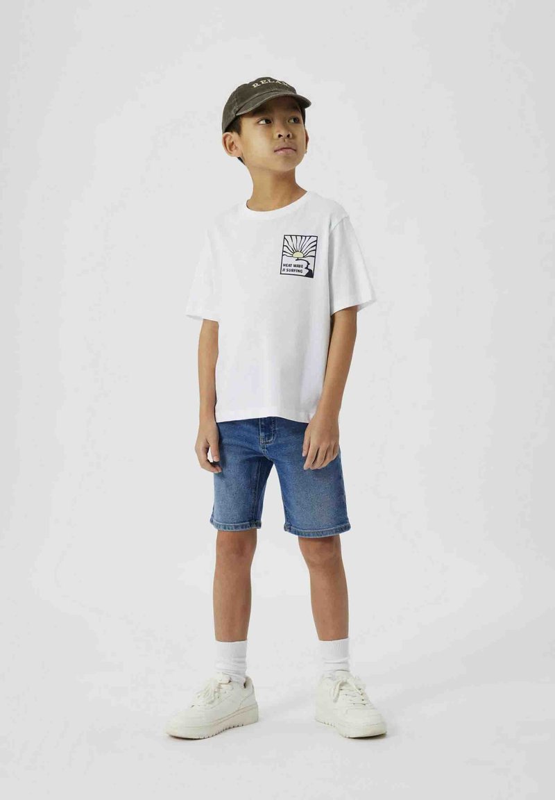 Camiseta blanca de algodón con un gráfico en el frente y pantalones cortos de mezclilla azul. El niño lleva zapatillas blancas y una gorra verde oscura. Fondo liso.