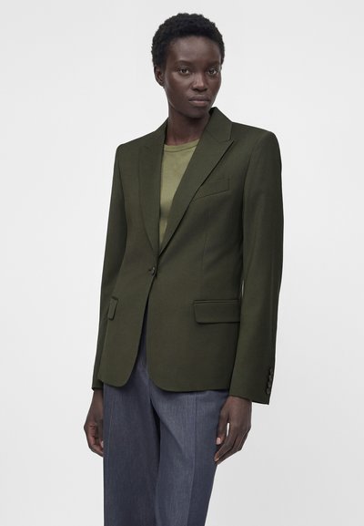Γυναίκα μοντέλο φοράει ένα σκούρο πράσινο σακάκι με ένα κουμπί πάνω από ένα ανοιχτό ελιά πουκάμισο και γκρι παντελόνια ραμμένα στα μέτρα της, σε απλό φόντο.