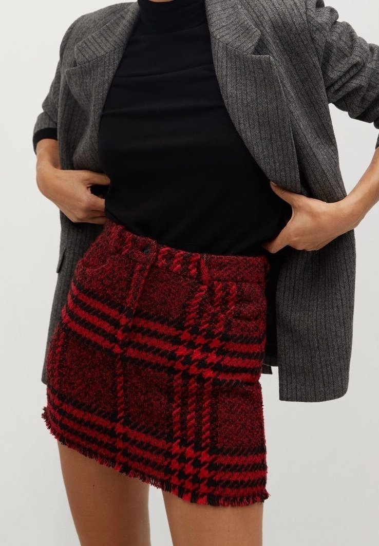 Femme portant un haut noir, un blazer gris à fines rayures, et une mini-jupe texturée à carreaux rouge et noir avec un ourlet effiloché.