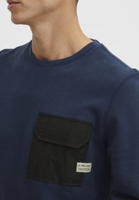 Marineblå hettegenser med en svart lomme med label. Myk tekstur, rund hals og synlige sømmer.