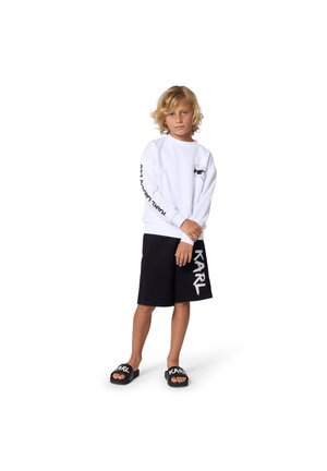 Sweat-shirt blanc à col rond avec un détail graphique noir sur la manche, associé à un short noir portant un grand logo blanc. Sandales noires.