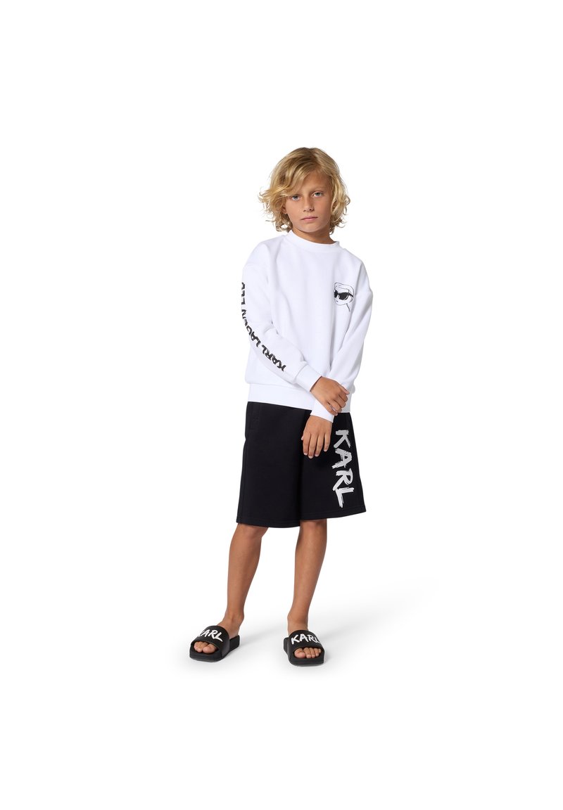 Λευκή φούτερ με στρογγυλή λαιμόκοψη και μαύρη γραφική λεπτομέρεια στο μανίκι, συνδυασμένη με μαύρο σορτς που έχει μεγάλο λευκό λογότυπο. Μαύρα σανδάλια.